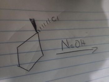 The image shows a chemical reaction diagram on lined paper. The diagram depicts a cyclopentane ring with a wedge line coming off one vertex, which is a representation of a chlorine (Cl) atom. 

The arrow pointing to the right indicates the reaction's progression and is labeled with "NaOH," representing sodium hydroxide as a reactant. This suggests a nucleophilic substitution reaction where NaOH is likely used to replace the chlorine atom with a hydroxyl group (-OH), converting the compound into an alcohol.

This type of reaction is common in organic chemistry where an alkyl halide is converted into an alcohol. The structure and notation exemplify the stereochemistry and orientation, with the wedge indicating that the chlorine atom is leaving the plane of the ring.